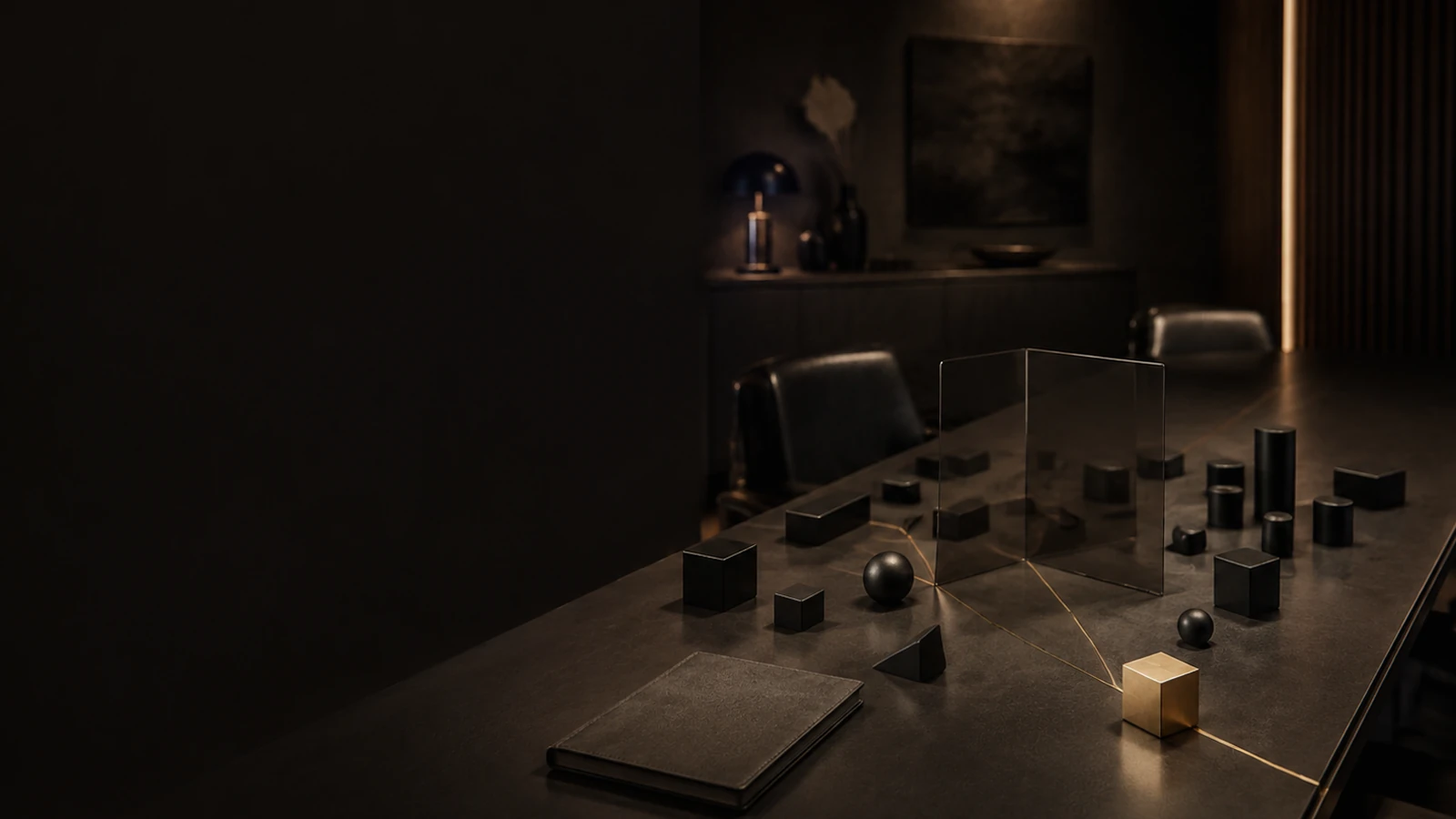 A dark boardroom table under a single lamp, set with small black blocks and spheres arranged around an upright glass panel, one brass cube among them, and a closed leather folio at the near edge.