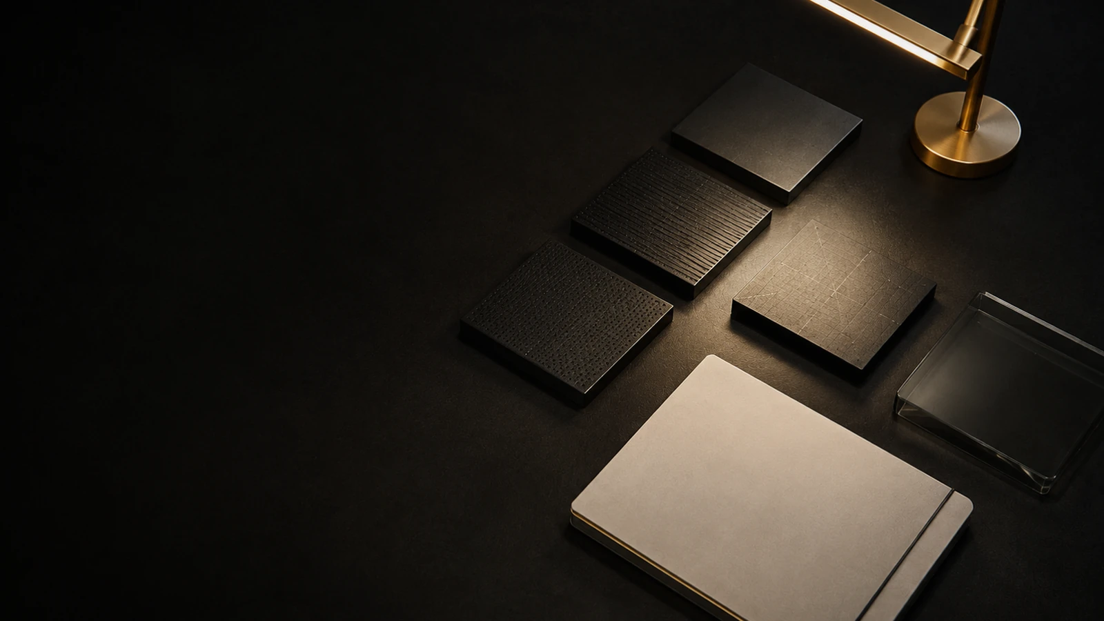 Four material squares in different surface textures fanned out under a brass desk lamp, with a cream notebook and a glass tray at the lower edge.