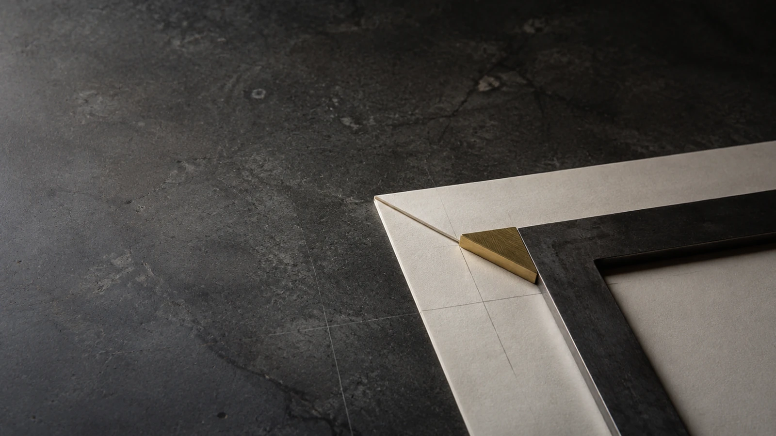 The corner of a pale mount board and a dark frame on concrete, joined by a small brass wedge over faint pencil register lines.