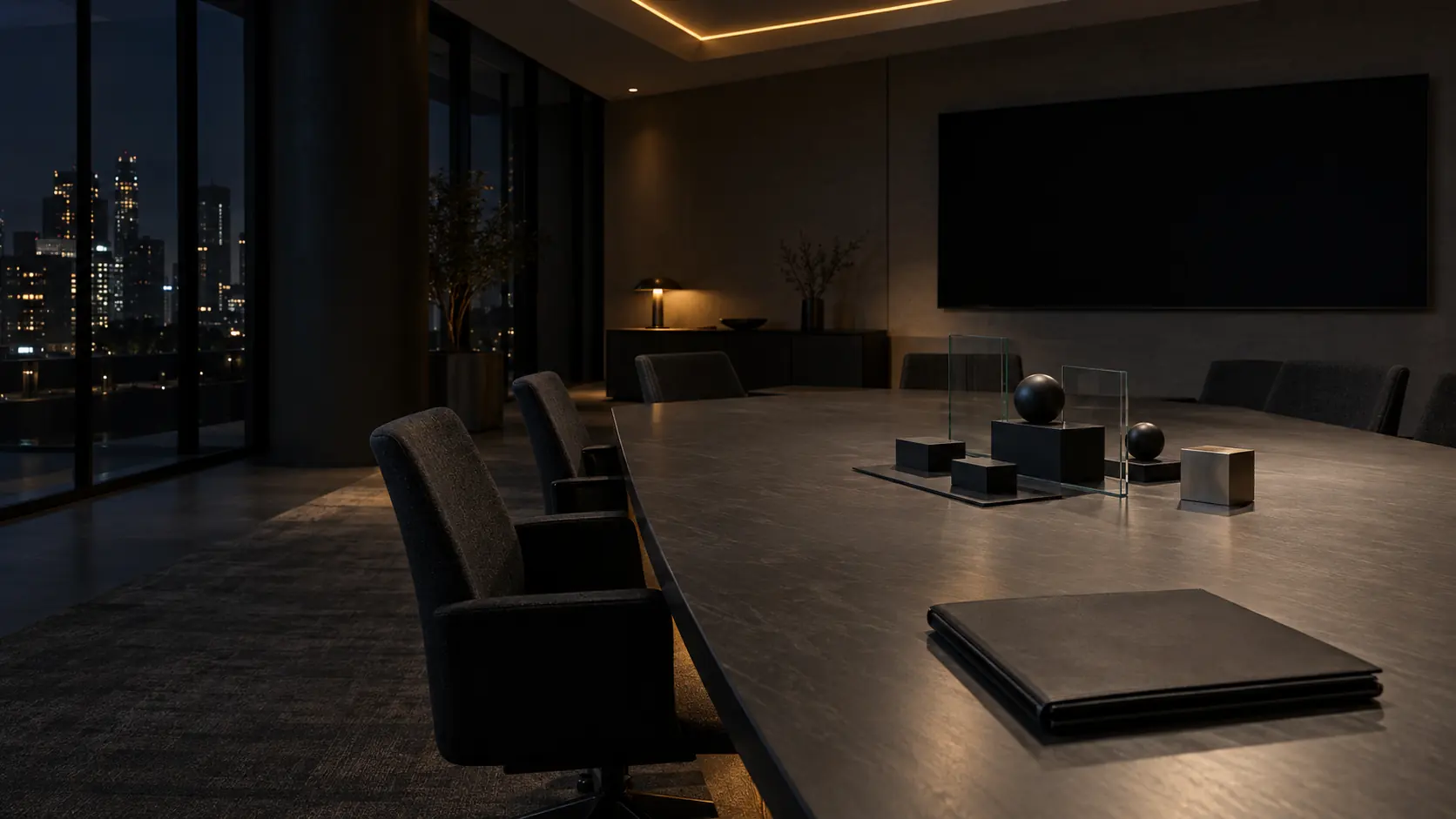 An empty boardroom at night: a long dark table under recessed lights, a closed folio at the near end, a switched-off wall screen and the city lit beyond the glass.
